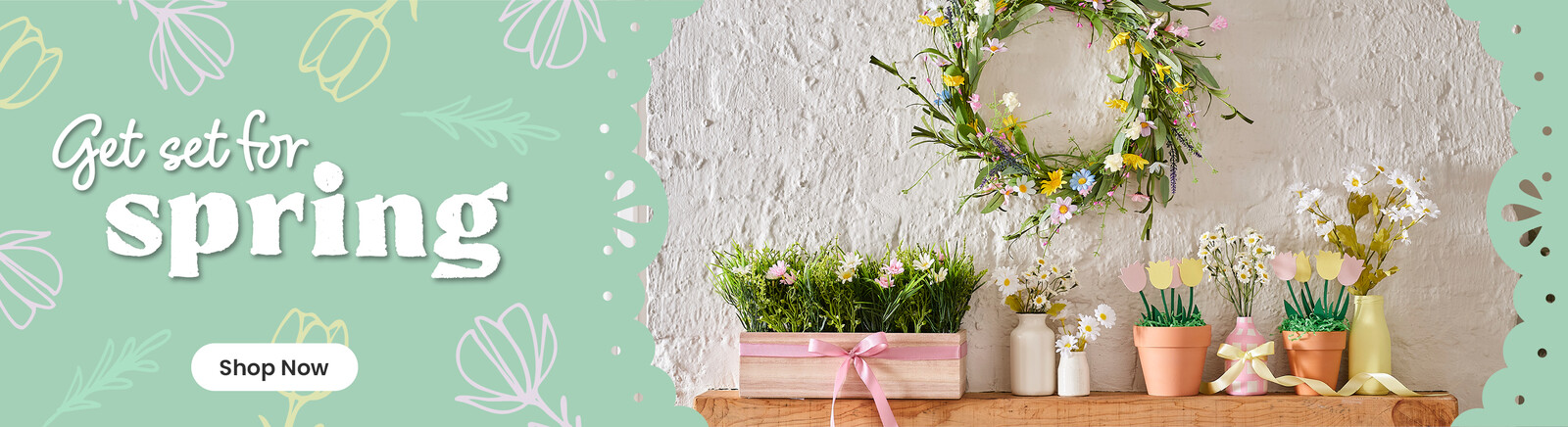 A spring wreath with pastel flowers on a wall, underneath on a shelf are vases with flowers and pots with paper flowers in them. Header text says Get set for Spring, along with a Shop Now button.